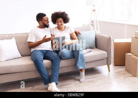 Happy Black Spouses Using Tablet Sitting Among Moving Boxes Indoor Stock Photo