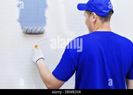Service Repair and restoration of premises. Portrait of a house painter and a paint roller painting the wall in blue. View from the back. A worker in Stock Photo
