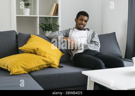 Cheerful curly haired ethnic african american male in casual clothes resting at home on comfortable sofa while watching tv channels or movie during we Stock Photo