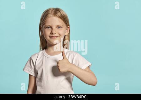 Close-up studio shot of a wonderful blonde little lady with a long hair, in a white t-shirt, showing thumb up and looking happy while posing over a bl Stock Photo