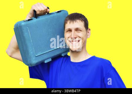 Portrait of a positive young repairman on a yellow background with a wide smile. A worker of Caucasian appearance holds a tool case on his shoulder. U Stock Photo