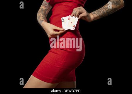 Slender woman with tattoos on arms in red cocktail dress standing against black background, holding pair of winning aces near waist, cropped shot. Pas Stock Photo