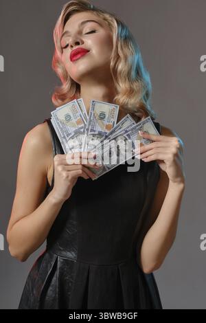 Gorgeous blonde woman with bright make-up, in black stylish dress is holding some money and posing against gray studio background. Gambling entertainm Stock Photo