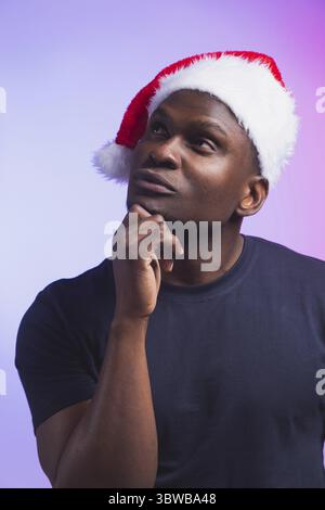Portrait of pensive african american man in santa hat and casual t-shirt on colourful background, christmas time. Winter holidays and emotional people Stock Photo