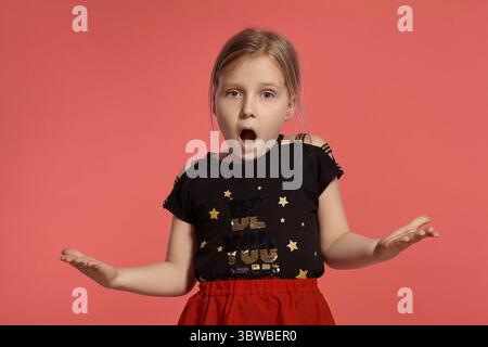 Close-up shot of a charming blonde little lady in a combined dress, looking shocked while posing against a pink background with copy space. Concept of Stock Photo
