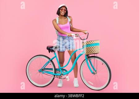 Positive young black lady in fashionable summer clothes posing with classic bicycle on pink studio background Stock Photo