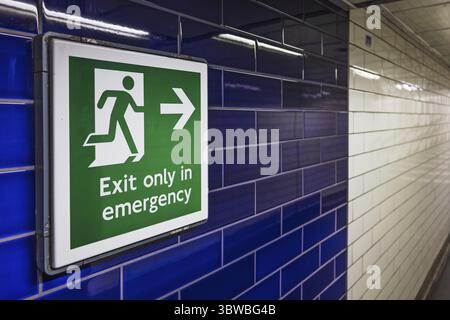 Green emergency exit sign with arrow on blue tiled subway wall in London Underground Stock Photo