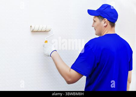 A painter in a cap and gloves paints the wall with a paint roller. Portrait of a young worker in uniform. Professional repair of premises. Mockup Stock Photo