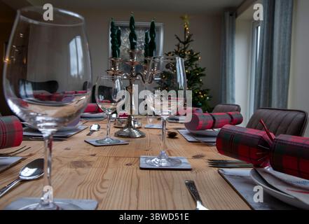 Festive dining table set with glassware and Christmas crackers in a cozy room with a decorated tree and candles Stock Photo