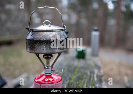 Worn metal kettle on gas stove atop weathered wooden table outdoors, blurred camping mug, thermos and forest path in background, focus on the kettle. Stock Photo