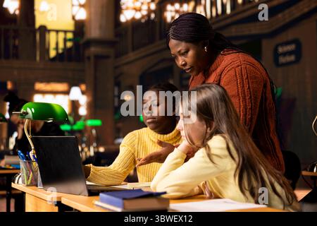 African american professor providing instruction to diverse girls during a private learning session, assisting with homework and school projects in a library. Doing tutoring activity for guidance. Stock Photo