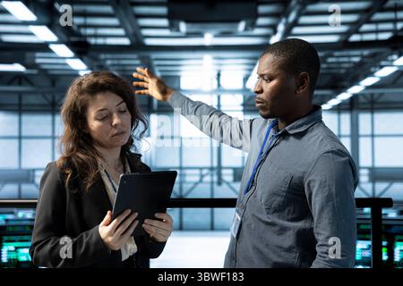 Teamworking admins in data center automating routine maintenance tasks with tablet, executing diagnostic scripts. Server farm colleagues using device, adjusting cooling and power settings Stock Photo