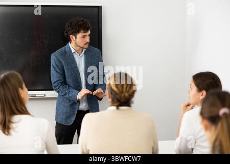 Male business coach standing near interactive board explaining topic while standing against group of people sitting at desks in front of him Stock Photo
