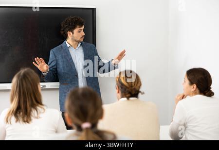 Male business coach standing near interactive board explaining topic while standing against group of people sitting at desks in front of him Stock Photo
