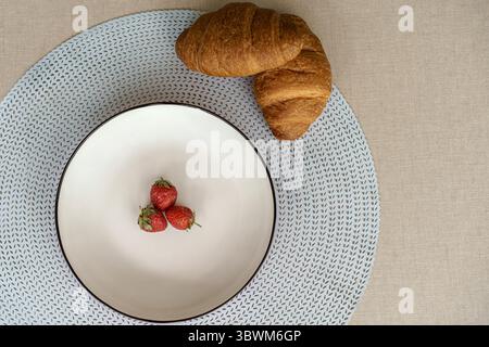 Minimalist breakfast flat lay. Top view of three fresh strawberries on a white plate with croissants on a blue placemat. Healthy eating concept. Stock Photo