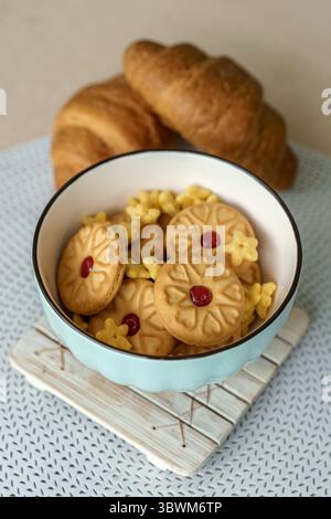 Teatime still life featuring a green bowl of jam-filled shortbread cookies and freshly baked croissants on a beige background. Stock Photo