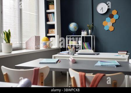 Empty classroom with desks and chairs arranged near window, school supplies including books, globe, and stationery on shelves, bright daylight illuminating modern educational space Stock Photo