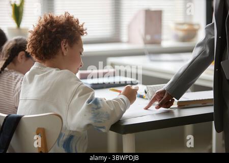 Caucasian boy sitting at desk writing while adult Black woman standing beside him pointing at paper, classroom setting with other children visible in background, learning activity Stock Photo