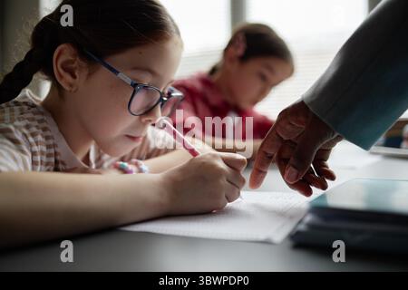 Caucasian girl child wearing glasses writing in notebook while hand of Black female teacher pointing at paper, another girl child sitting in background focusing on schoolwork in classroom setting Stock Photo