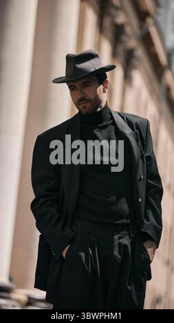 Man in black suit and fedora hat posing outdoors with columns in background Stock Photo
