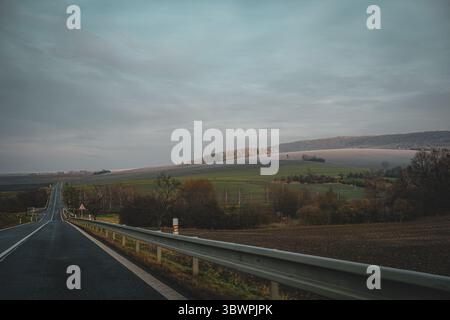 Empty countryside road with guardrail leads through rolling fields toward frost-covered hills under a cloudy sky. Stock Photo