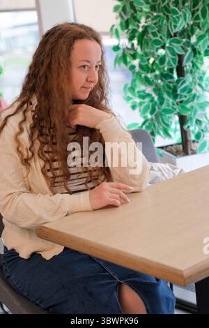 A young woman in a cozy sweater sits at a cafe table, deep in thought. Stock Photo