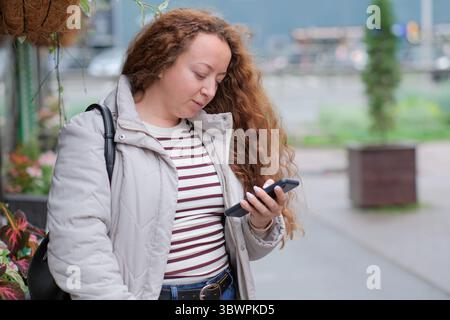 A young woman stands on a colorful street, engaged with her phone outdoors. Stock Photo