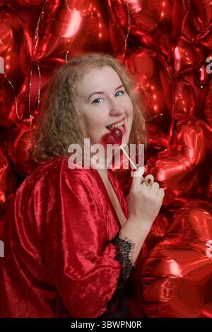 An older pretty blonde curly woman in a playful red robe poses in the studio with heart-shaped balloons. Stock Photo