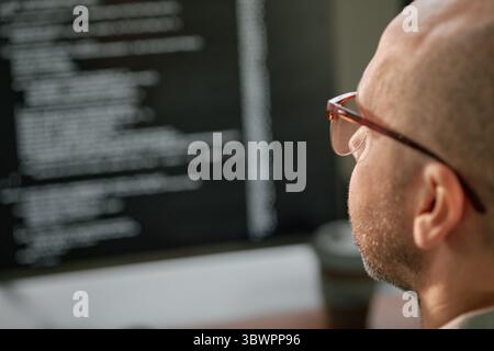 Caucasian middle aged man wearing glasses working on computer monitor displaying lines of code, sitting at desk in office environment, focusing on programming task Stock Photo