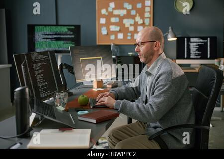 Middle aged Caucasian man working at computer desk in modern office, typing on keyboard while analyzing code on multiple monitors, wearing glasses and focused on programming tasks Stock Photo