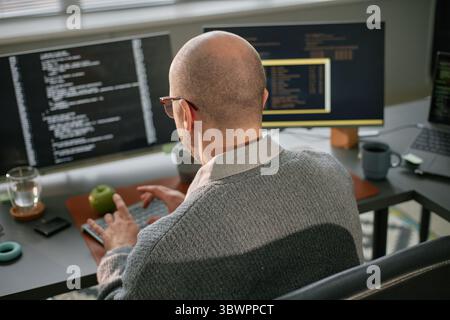 Middle aged Caucasian man sitting at desk working on computer with multiple monitors displaying programming code, typing on keyboard with glass of water and green apple nearby Stock Photo