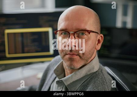 Portrait of middle aged Caucasian man wearing glasses looking at camera while sitting at desk with computer monitors displaying programming code in modern office setting Stock Photo