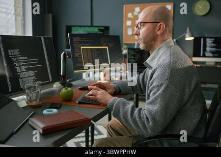 Middle aged Caucasian man working at computer desk, typing on keyboard and analyzing code on multiple monitors, sitting in modern office environment with digital equipment Stock Photo