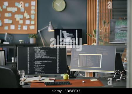 Modern office workspace featuring dual monitors displaying programming code, desktop computer, keyboard, notebook, green apple, glass of water, and blurred background with bulletin board Stock Photo