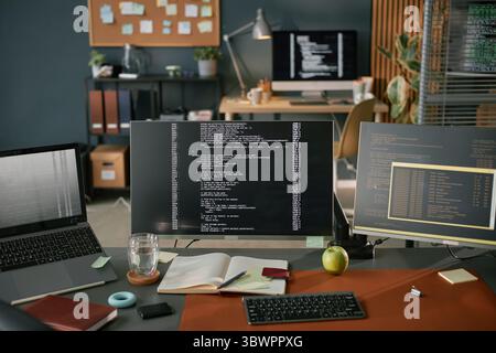 Three computer monitors displaying programming code on desk with open notebook, pen, glass of water, apple, and laptop, modern workspace suggesting software development environment Stock Photo