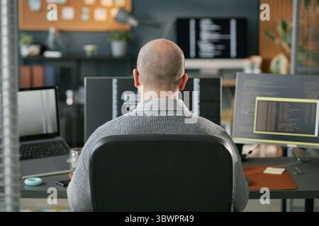 Middle aged Caucasian man sitting at desk working on multiple computer monitors with programming code visible on screens in modern office environment, back view Stock Photo