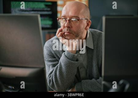 Middle aged Caucasian man wearing glasses sitting at desk working on computer monitors, resting chin on hand, appearing focused while looking at screen in office setting Stock Photo