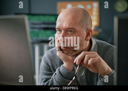 Caucasian middle aged man sitting at desk holding eyeglasses looking at computer monitor with thoughtful expression hand resting on face working in modern office environment Stock Photo