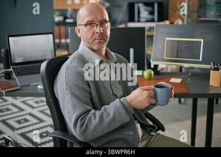 Portrait of middle aged Caucasian man with glasses sitting in modern office holding coffee mug looking at camera, computer monitors with code visible in background Stock Photo