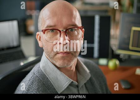 Portrait of middle aged Caucasian man with glasses looking into camera sitting at desk in modern office environment surrounded by computer monitors and technology equipment Stock Photo