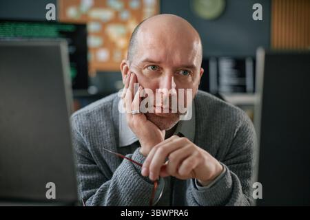 Portrait of middle aged Caucasian man resting chin on hand while sitting at desk, gazing intently at computer monitors, holding eyeglasses, appearing focused on work Stock Photo