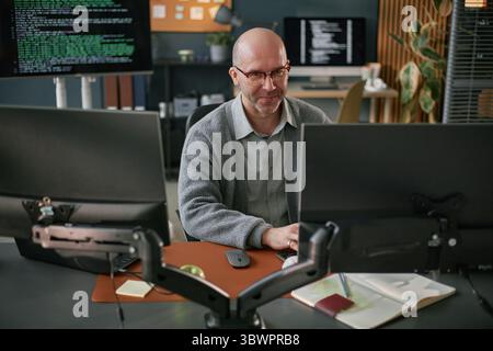 Middle aged Caucasian man sitting at desk working on computer monitors in modern office, typing on keyboard with programming code visible on screens in background Stock Photo