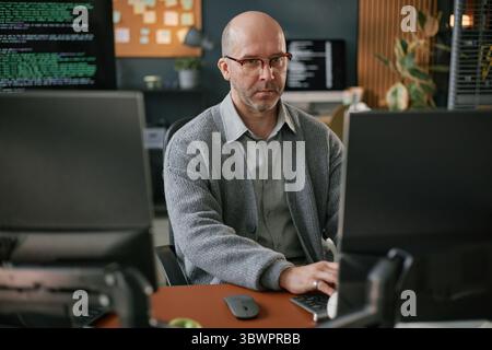 Portrait of middle aged Caucasian man wearing glasses sitting at desk working on computer monitors in modern office environment with programming code visible on screen Stock Photo