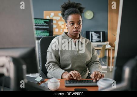 Young adult Black woman working at computer desk, focusing on typing with hands on keyboard, sitting in modern office environment with multiple monitors and programming code visible Stock Photo