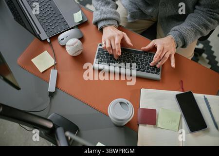 Caucasian middle aged man working at desk using wireless keyboard with laptop, smartphone, coffee cup and baseball on table, hands typing, workspace organized with sticky notes Stock Photo
