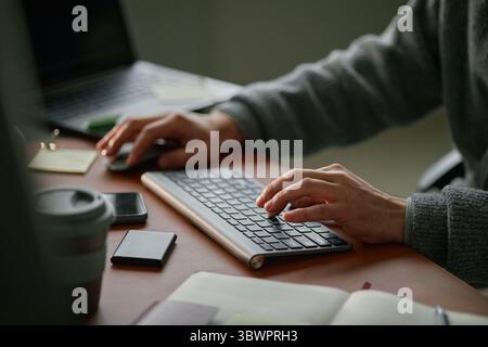Caucasian young adult man working at desk using wireless keyboard and mouse, hands typing on keyboard with smartphone, notebook, coffee cup and laptop visible on workspace Stock Photo