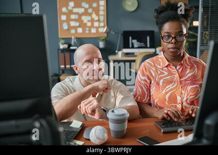 Middle aged Caucasian man and young adult Black woman working together at computer in modern office, both focused on screen, collaborating on project, coffee cup and baseball on desk Stock Photo