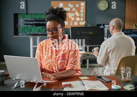 Black middle aged woman working on laptop at desk in modern office, typing and focusing on screen, Caucasian middle aged man sitting at workstation with computer monitors in background Stock Photo