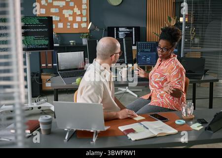 Black woman discussing project with Caucasian middle aged man in modern office, multiple computer monitors displaying code and data, collaborating on software development Stock Photo