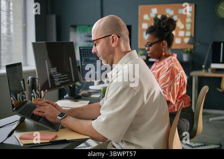 Middle aged Caucasian man typing on laptop at desk with large monitor displaying code, Black woman with glasses sitting behind working on computer in modern office workspace Stock Photo